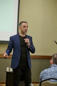 A man in bleu suit and black tee-shirt, giving a presentation